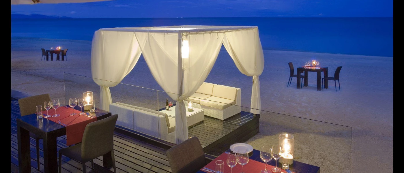 Wooden terrace with tables lit by lanterns, with the beach in front and a four-poster cabana with white drapes