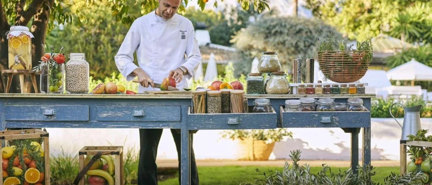 Staff member in white uniform cuts fruit on a table in a garden, surrounded by jars of grains and cereal