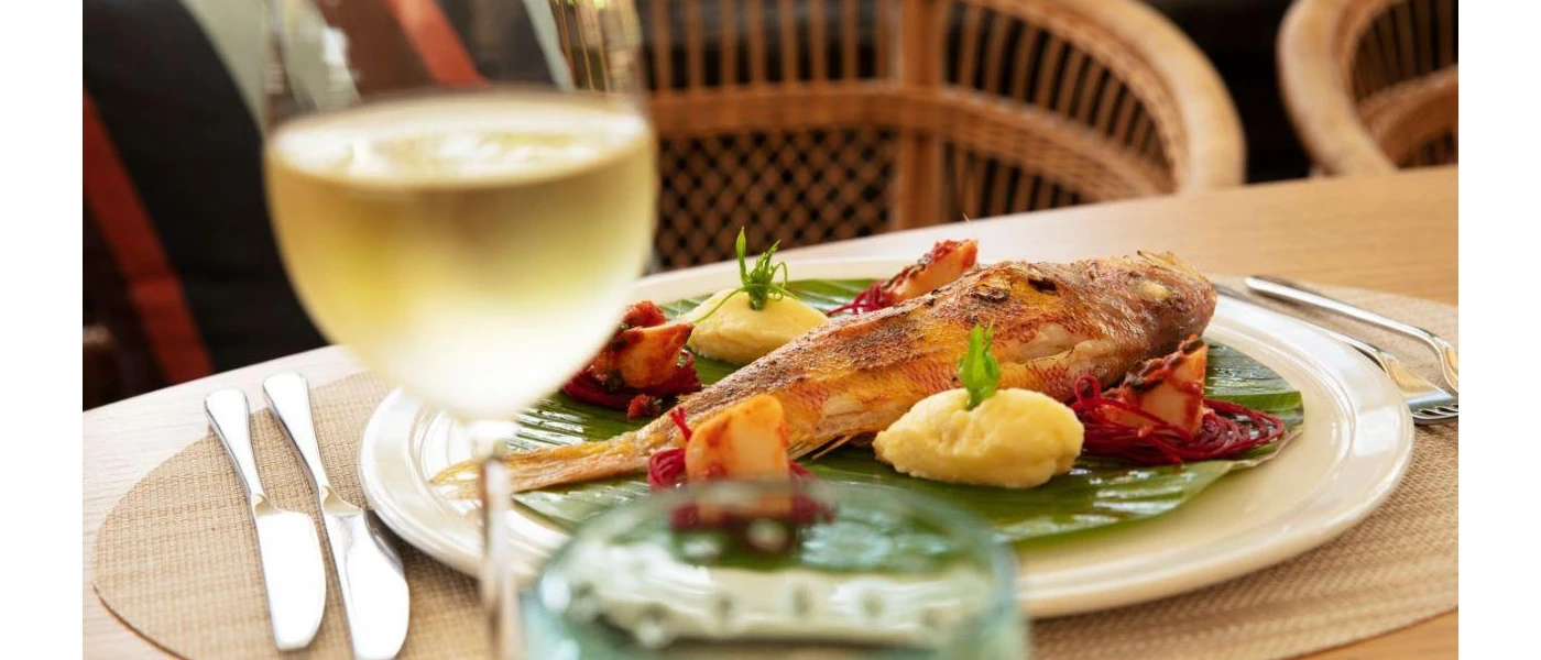 A plate of fish, mashed potato and vegetables on top of a bamboo leaf, next to a glass of crisp white wine