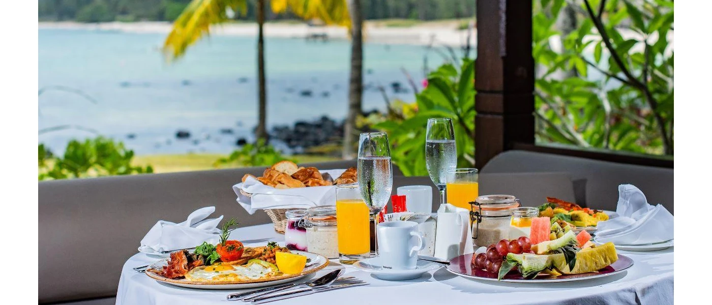 Table laid for breakfast, with two plates of eggs, beans, tomato and bacon, a basket of bread, a bowl of fruit and two glasses of orange juice, with the ocean in the background
