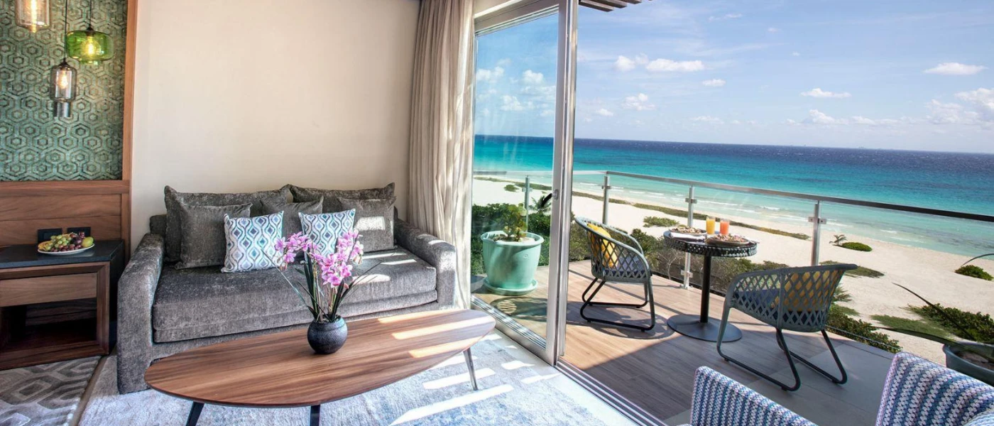 Hotel room seating area with a grey sofa, blue rug and wooden coffee table, next to an open furnished balcony overlooking the beach