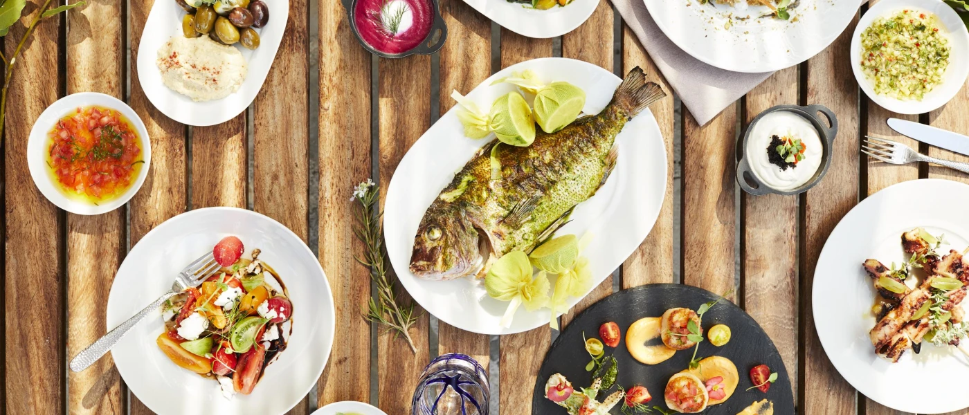 Wooden table filled with nutritious plates of food, including a grilled fish with limes, vegetable salads, grains and olives, and pots of dip
