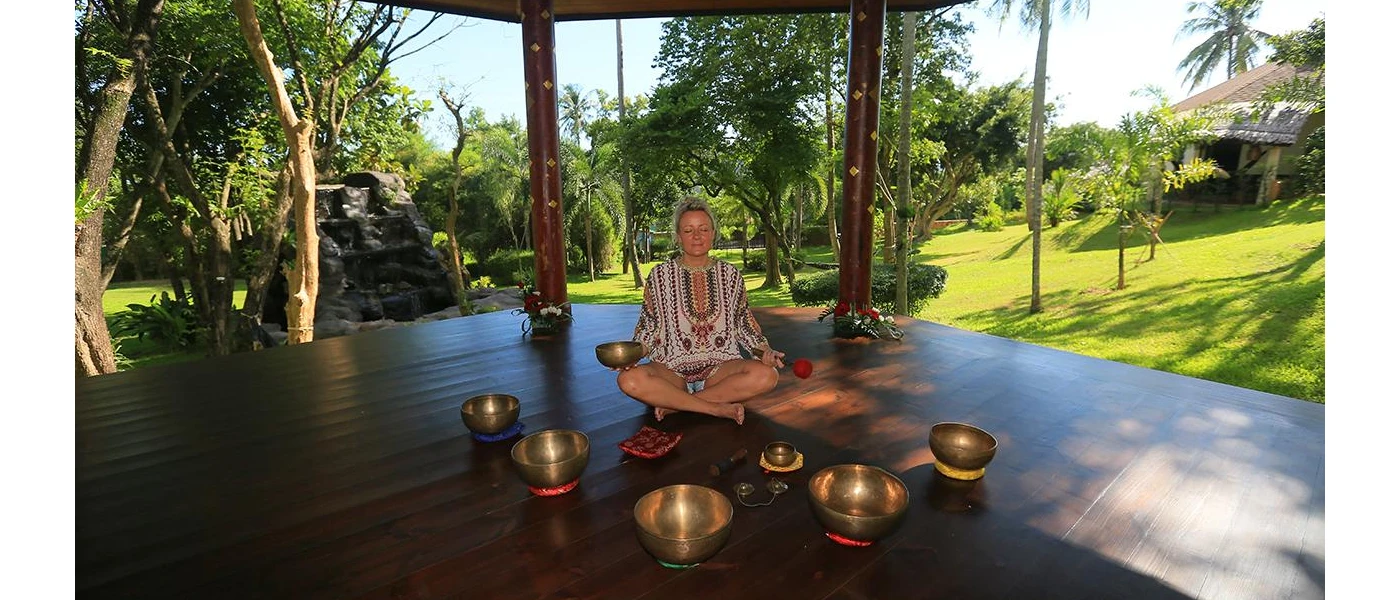 Smiling women with her eyes closed sits among sound bowls cross-legged in an open-air pavilion surrounded by gardens