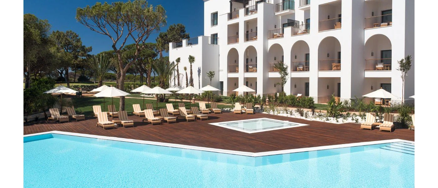 Multi-storey white building with an outdoor rectangular pool, smaller whirlpool set in wooden decking, striped loungers and white parasols under a blue sky