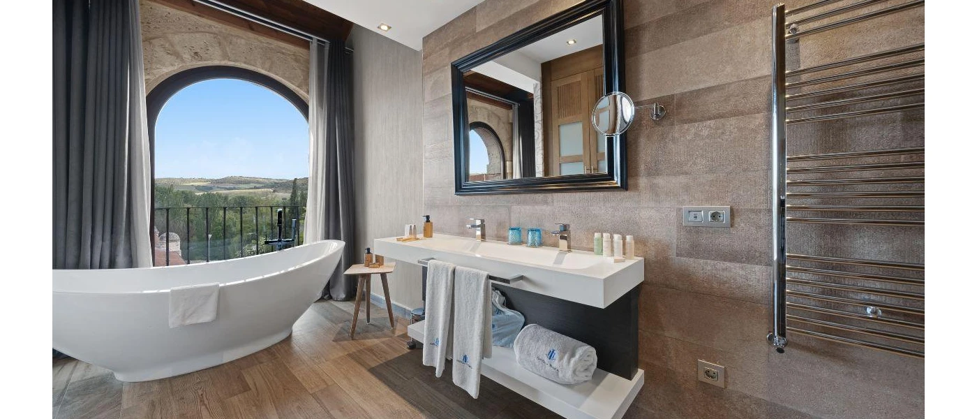 Bathroom with tiled walls and a wooden floor, a standalone bathtub and a view of the countryside from an arched window