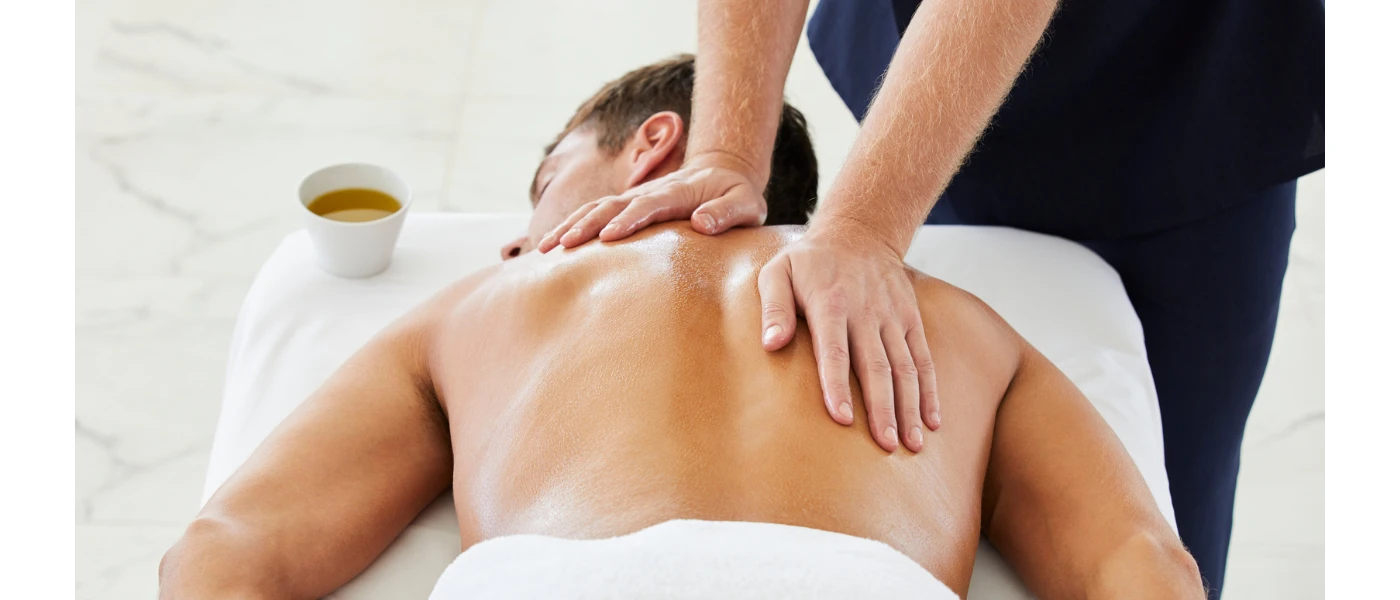 Man lies on his front in a towel as a therapist massages him with a pot of oil beside them