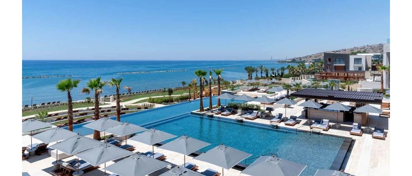 T-shaped swimming pool overlooking the ocean, surrounded by tall palm trees and blue-cushioned loungers shaded by blue umbrellas