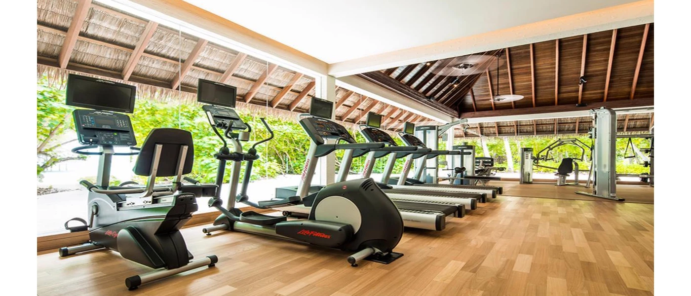 Treadmills and bikes lined up in a gym with wooden flooring, floor-to-ceiling windows and tropical garden views