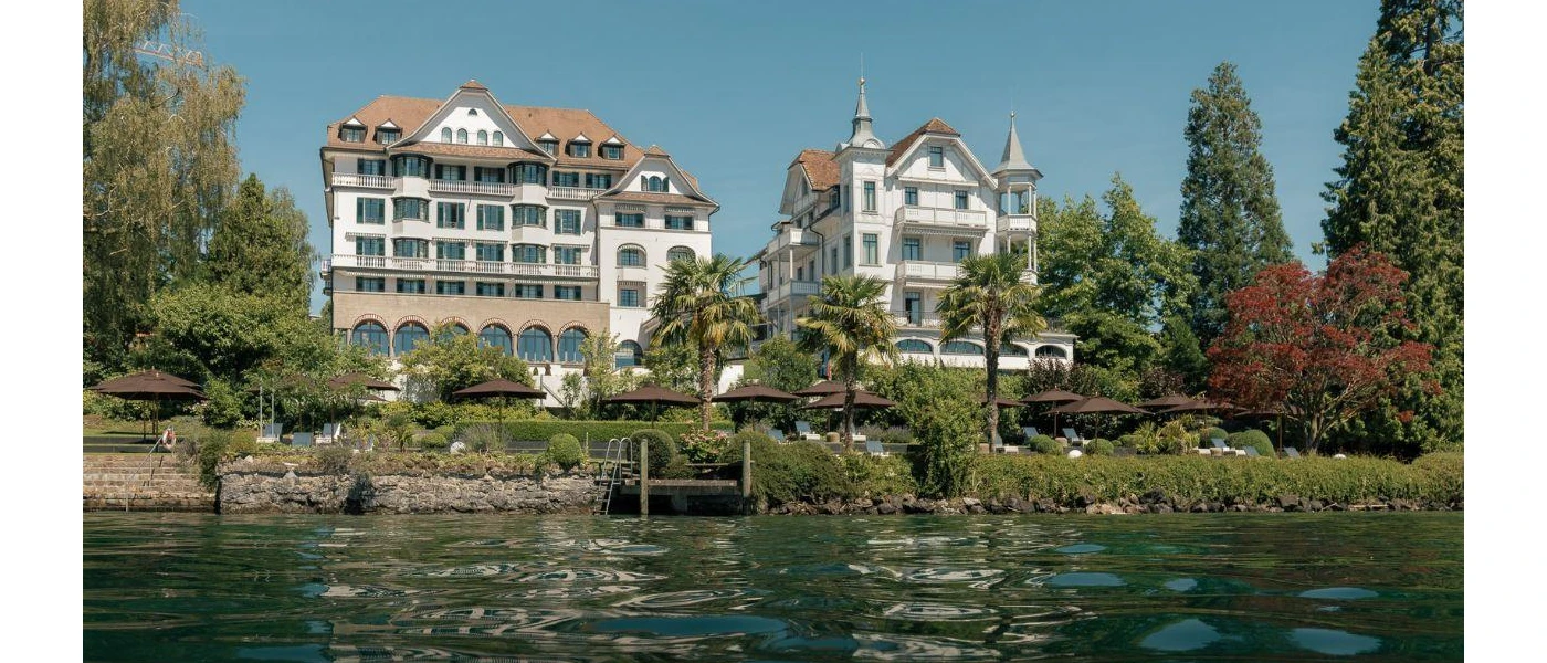 Lakeside view of Chenot Palace Weggin, fronted by palm trees shrubs and grassy lawns