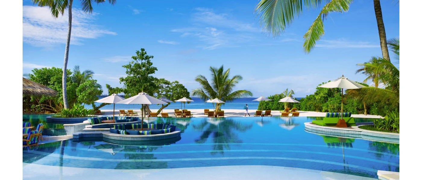 Palm-fringed swimming pool with cut outs for seating looking over white parasols, bright white sands and the ocean