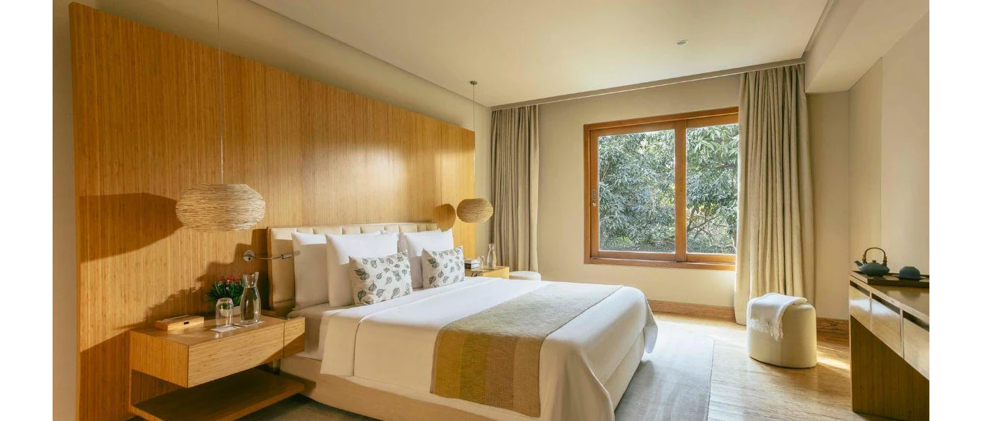 Neutral hotel bedroom with a wood-panelled back wall, white bedding, rattan hanging lights and a window with a view of greenery
