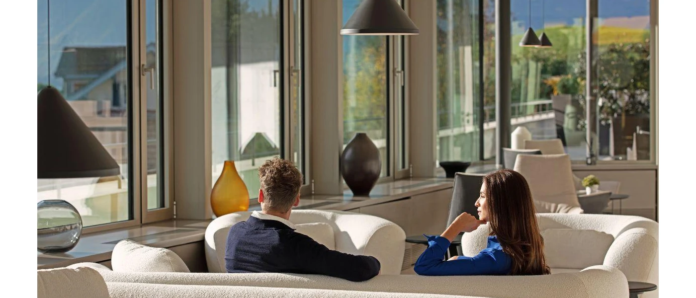 Man and woman sit in ivory seating in a sunny chic room with glass vases and floor-to-ceiling windows 