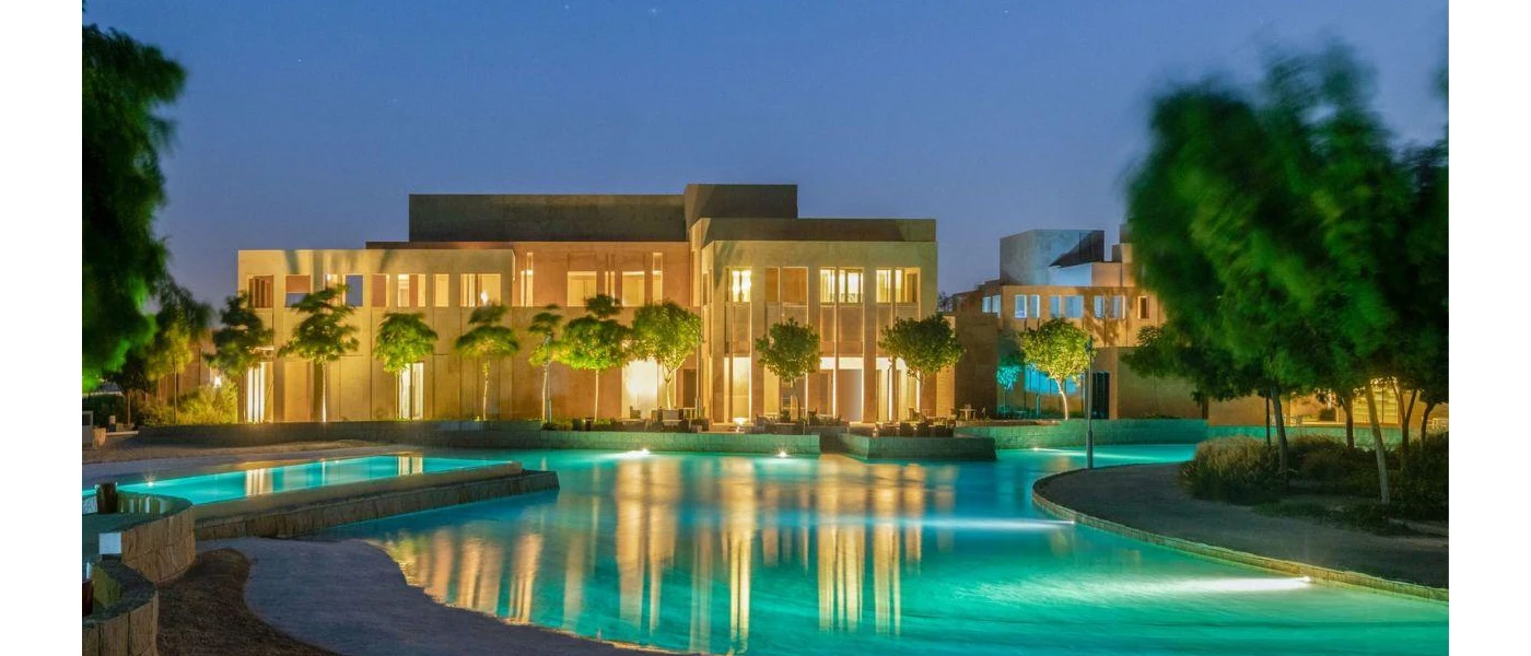 Cube-shaped sand-coloured building with a swimming pool in front lit up under a darkening sky