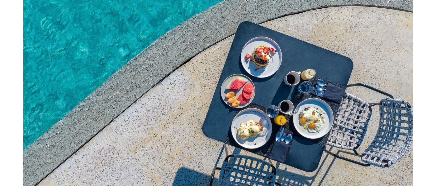 Poolside table set for breakfast, with plates of watermelon, fried and poached eggs, and a pancake stack topped with blueberries and strawberries