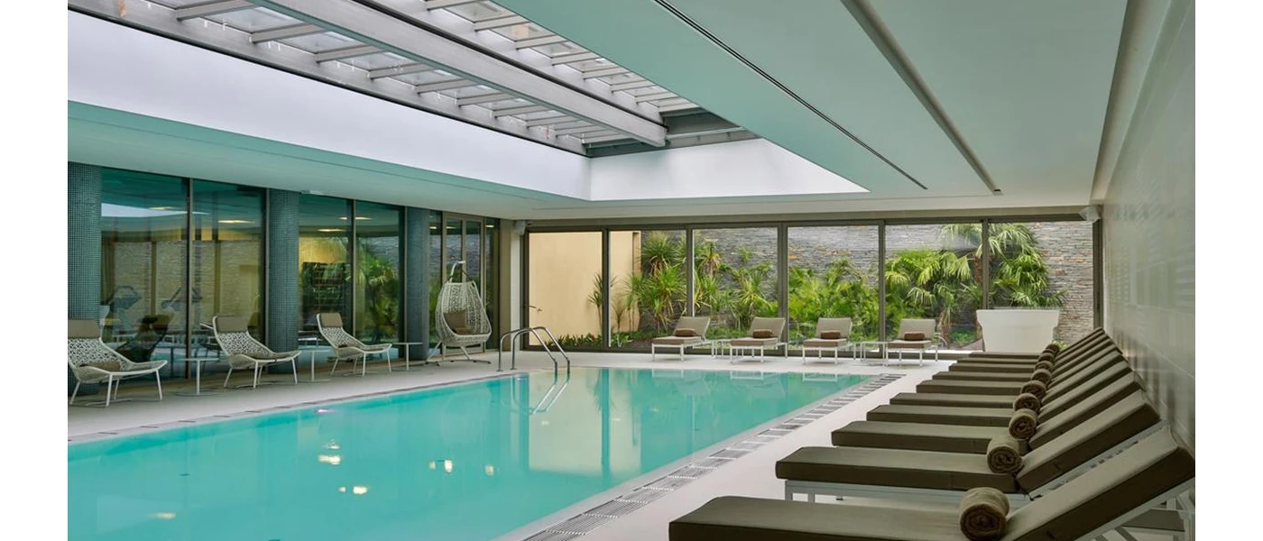 Rectangular indoor pool with floor-to-ceiling windows and grey-cushioned loungers lined up along the right-hand side