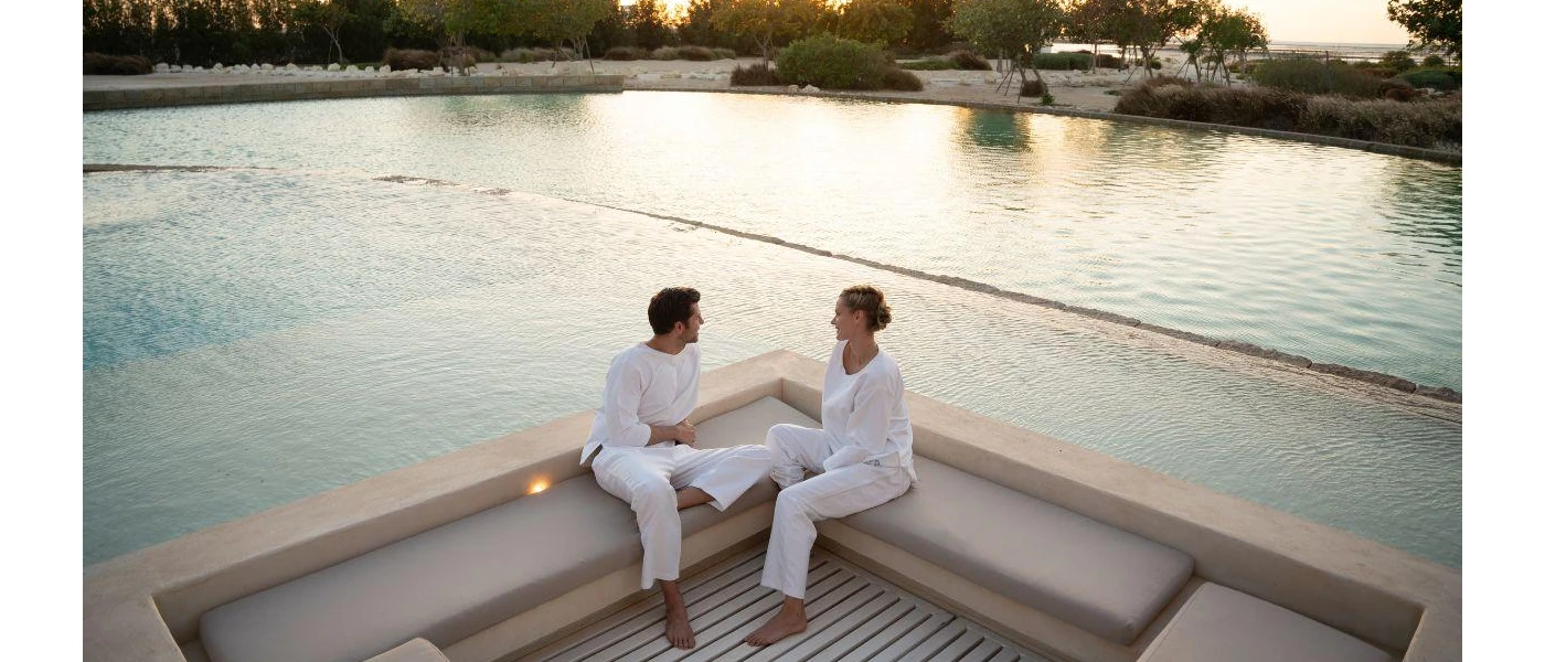 Man and woman in white relax on a neutral sofa next to a pool and beach as the sun sets behind them