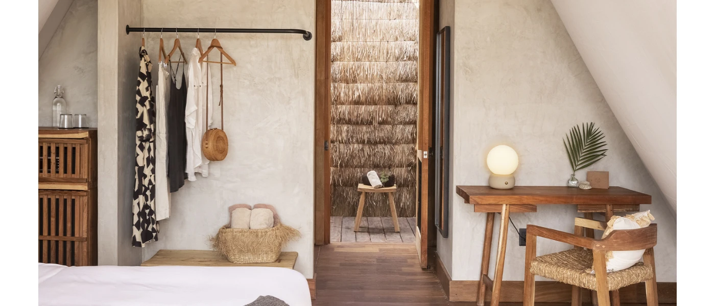 Room with white walls and flooring, a wooden desk area, wardrobe space with black and white clothes and rattan handbags, and an outdoor shower showing through a doorway