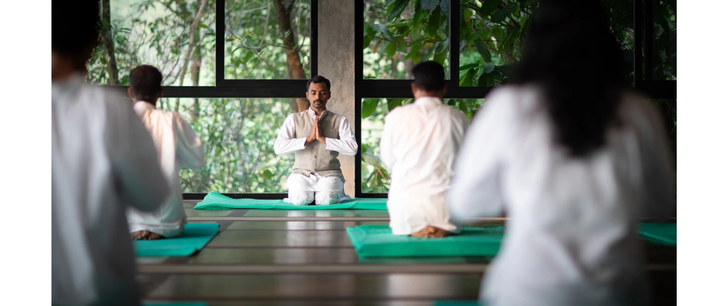 Group in white sit with their hands clasped in prayer on green yoga mats in a wooden studio