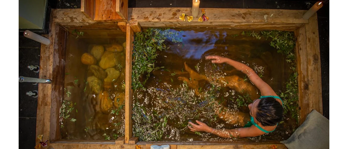 Woman in a blue swimsuit in a wooden bath with stones at the bottom and herbs in the water