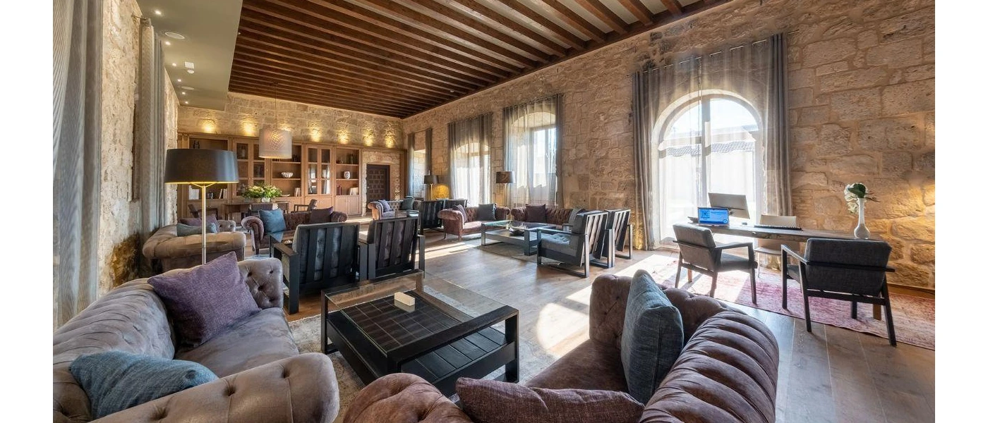 Lounge area with grey sofas, stone walls, large arched windows with sheer curtains, and a wooden cabinet containing books and ornaments