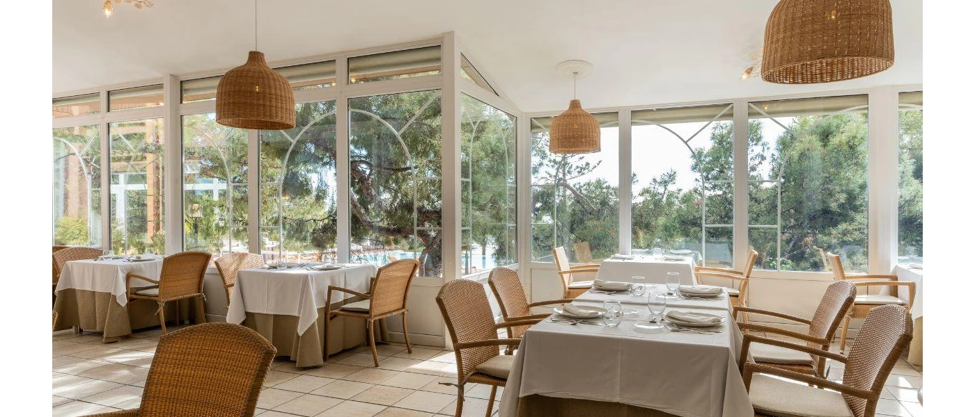 Airy restaurant with large wraparound windows, tables with white tablecloths and wooden chairs, and rattan pendant lights hanging from the ceiling