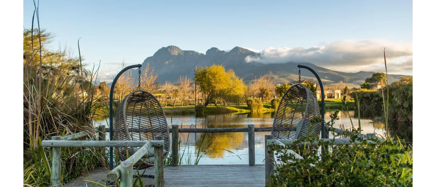 A lake framed by greenery and mountains, with a wooden deck and egg-shaped wicker chairs for enjoying the view