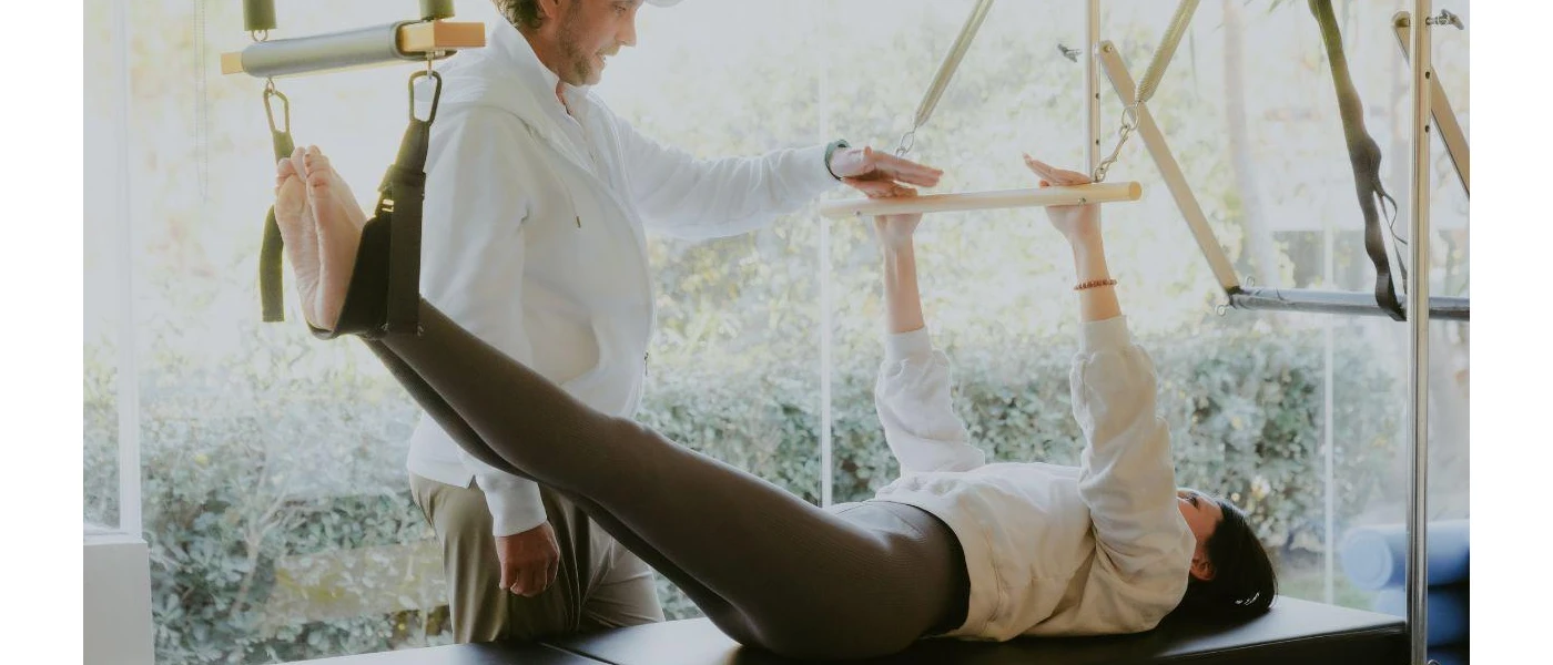 Woman lies on her back with her arms and feet outstretched in a reformer Pilates machine, as an expert instructor guides her