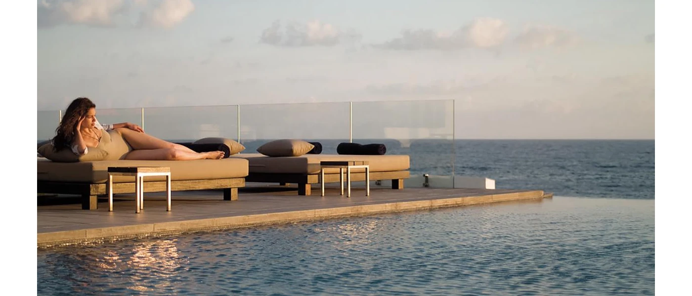 Woman relaxing on a cushioned lounger next to an infinity pool with a glass-walled edge and a sea view