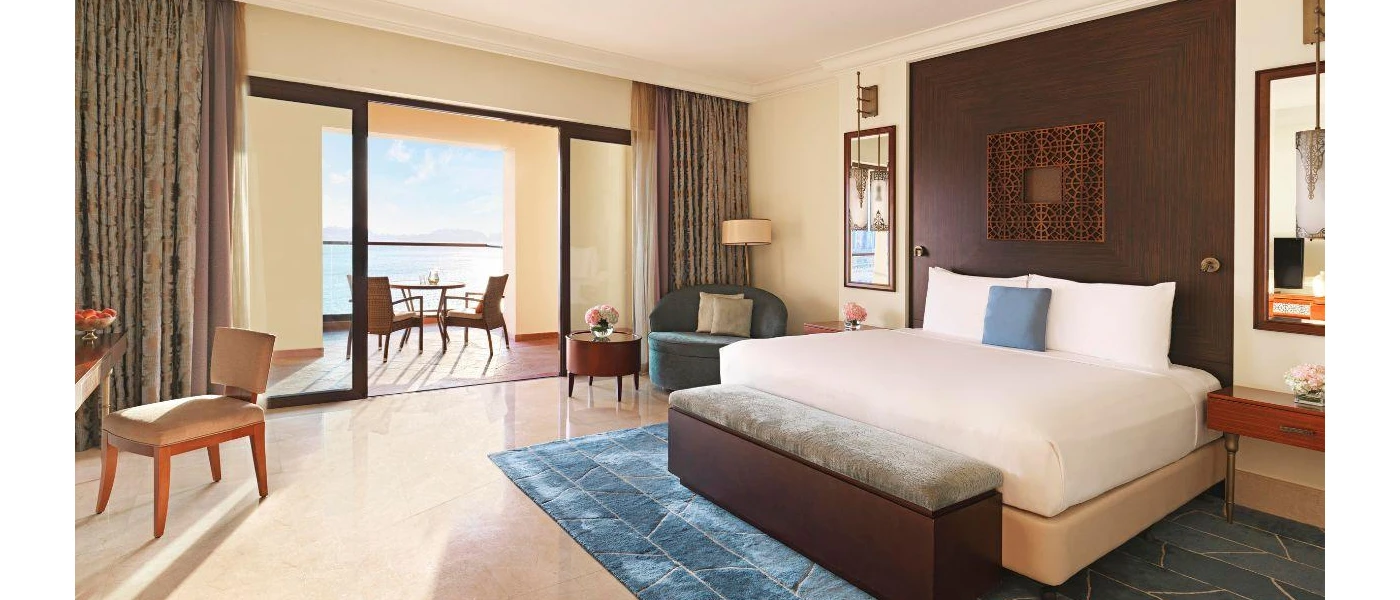 Hotel room with a dark-wood headboard, bench and coffee table, a blue rug, armchair and cushion, smooth tiled floors and giant patio doors opening onto a balcony