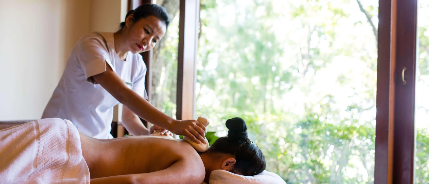 Woman lying down partially covered by a white towel, with her face in a massage bed hole, being massaged by a therapist in front of a window overlooking the forest