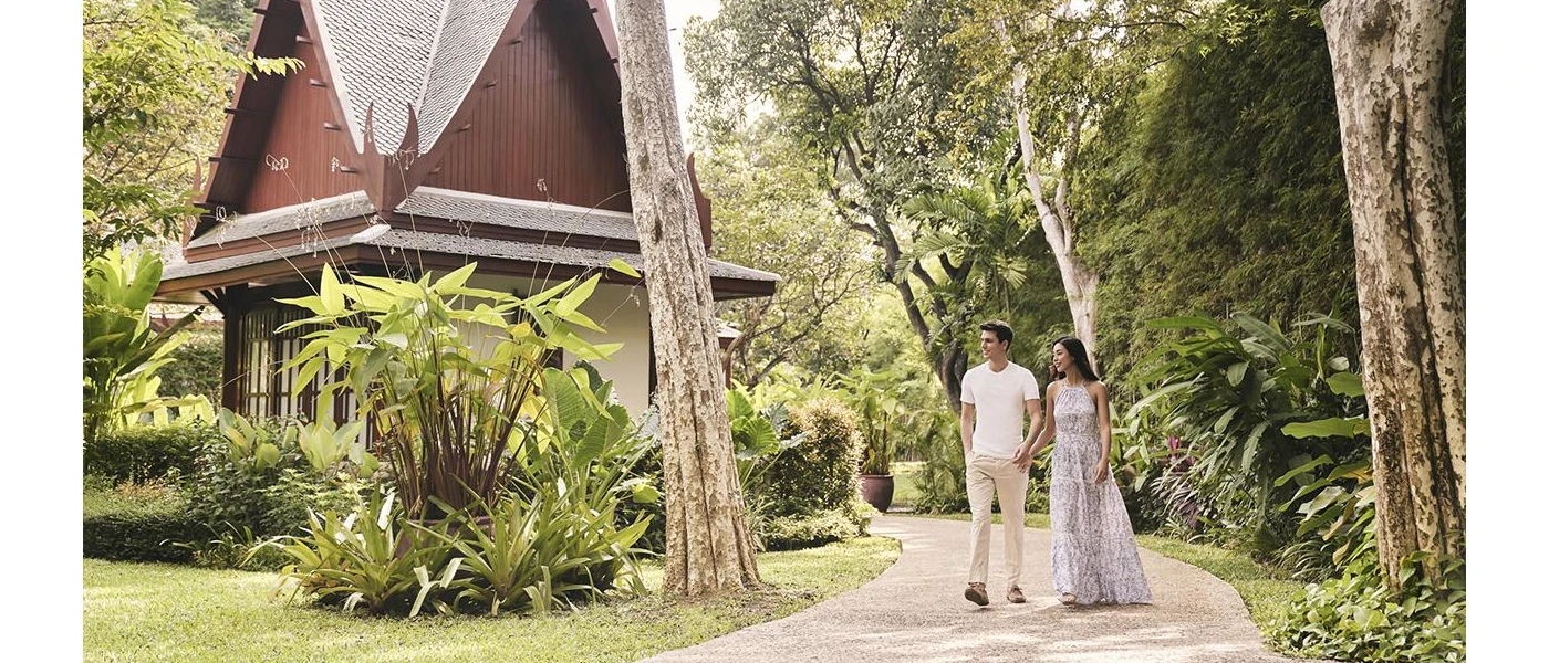 Man and woman stroll through tropical gardens hand in hand, with a pavilion to one side