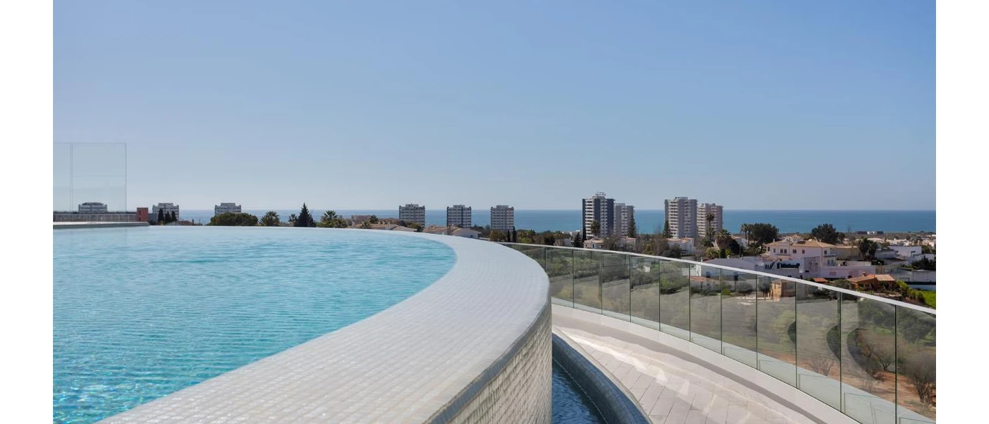 Swimming pool with a curved infinity edge, overlooking a glass balcony and a high-rise seafront town in the distance