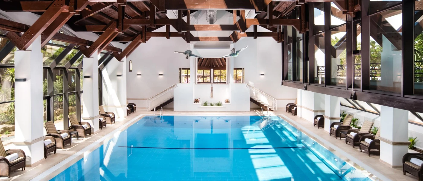 Rectangular indoor pool with a dark wood-beamed ceiling and armchair-style seating lined up at the water's edge