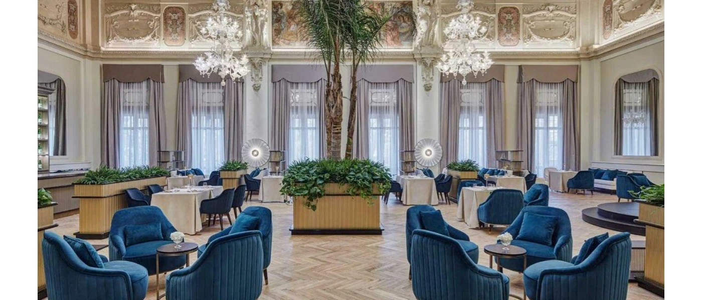 High-ceiling room with chandeliers, a central palm tree and tables with white clothes and blue velvet chairs