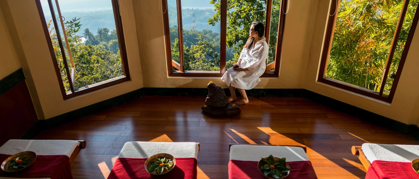 Woman in a white robe in front of a row of massage beds, sitting by a window with a view of the green mountains outside