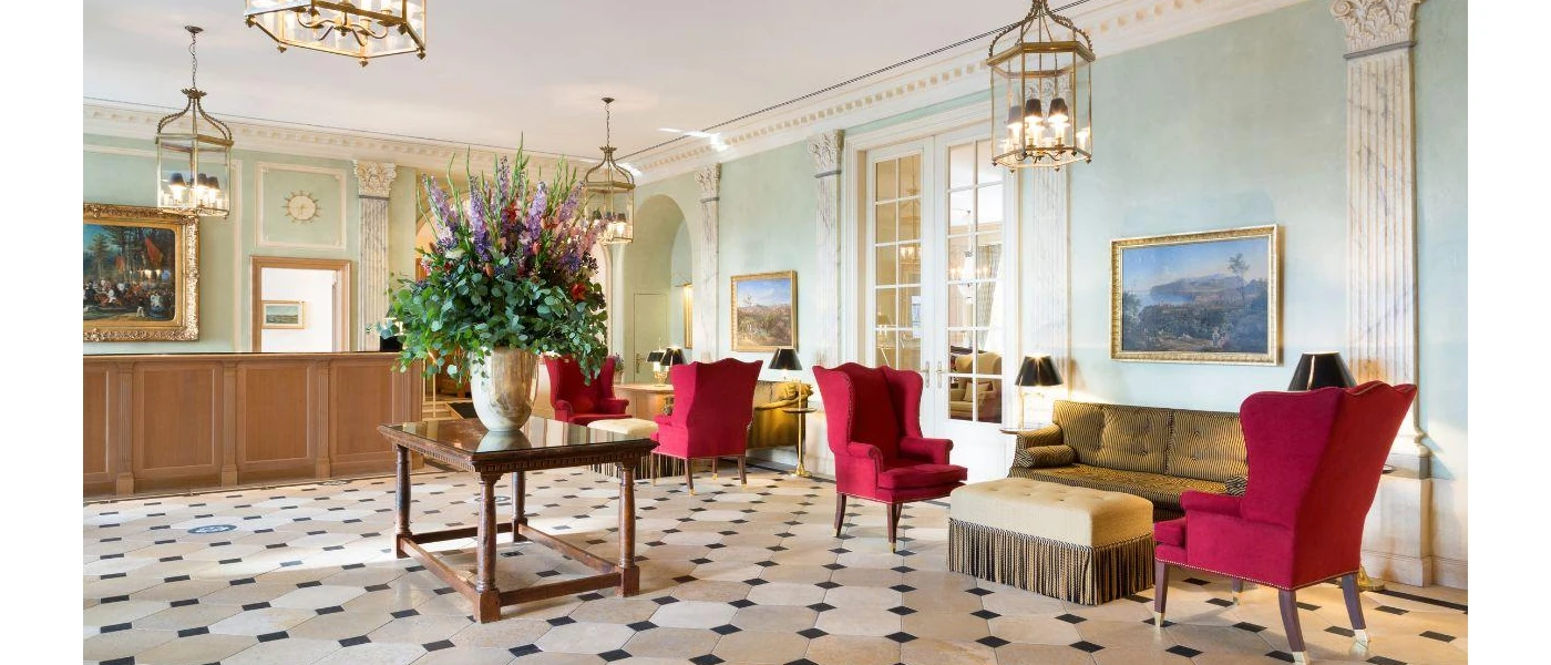 Elegant reception area with red velvet armchairs, a chequered floor, table with large flower arrangement, and wooden desk with chandelier-style lighting