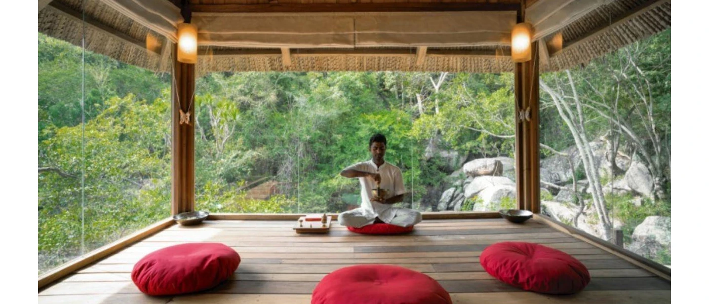 A man sits cross-legged in a yoga shala with panoramic windows, with three red cushions laid out ready to welcome guests