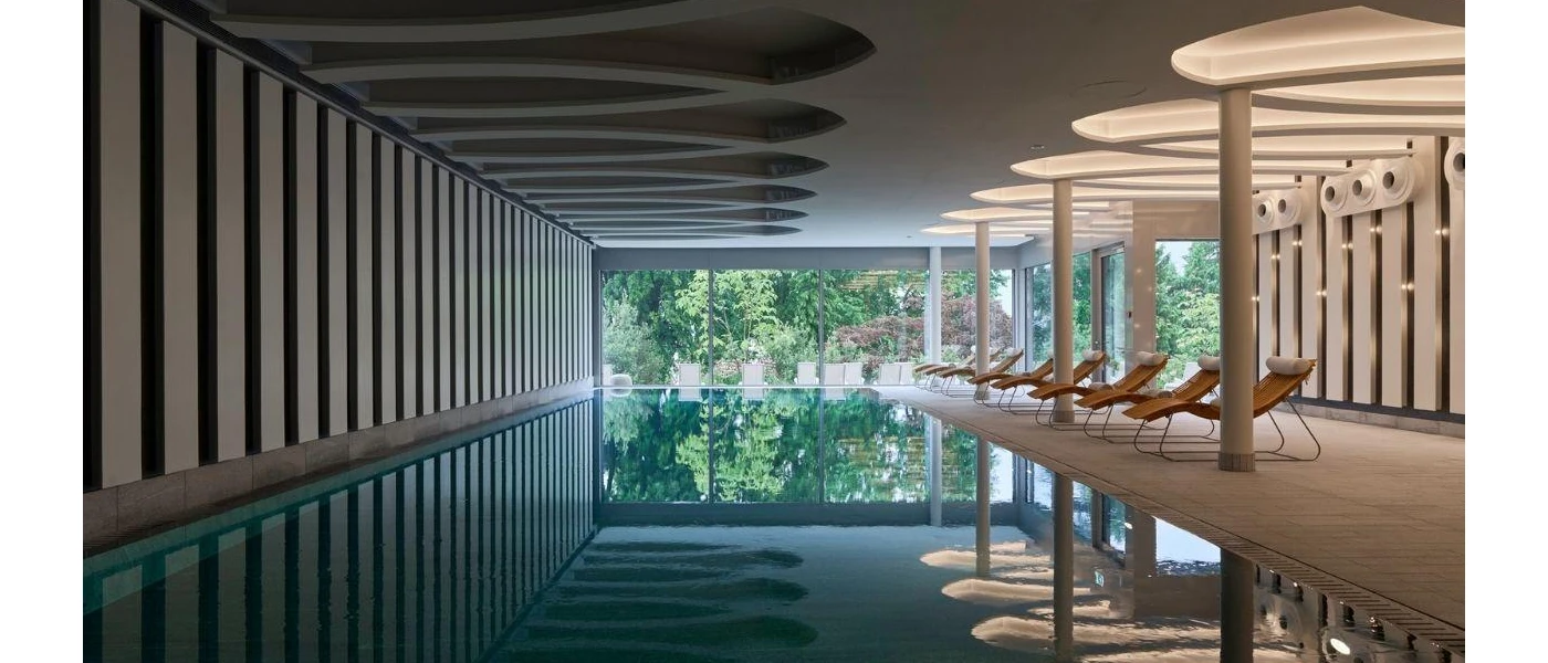 Indoor pool area with wooden loungers, mood lighting and full-length windows with a view of tropical greenery outside