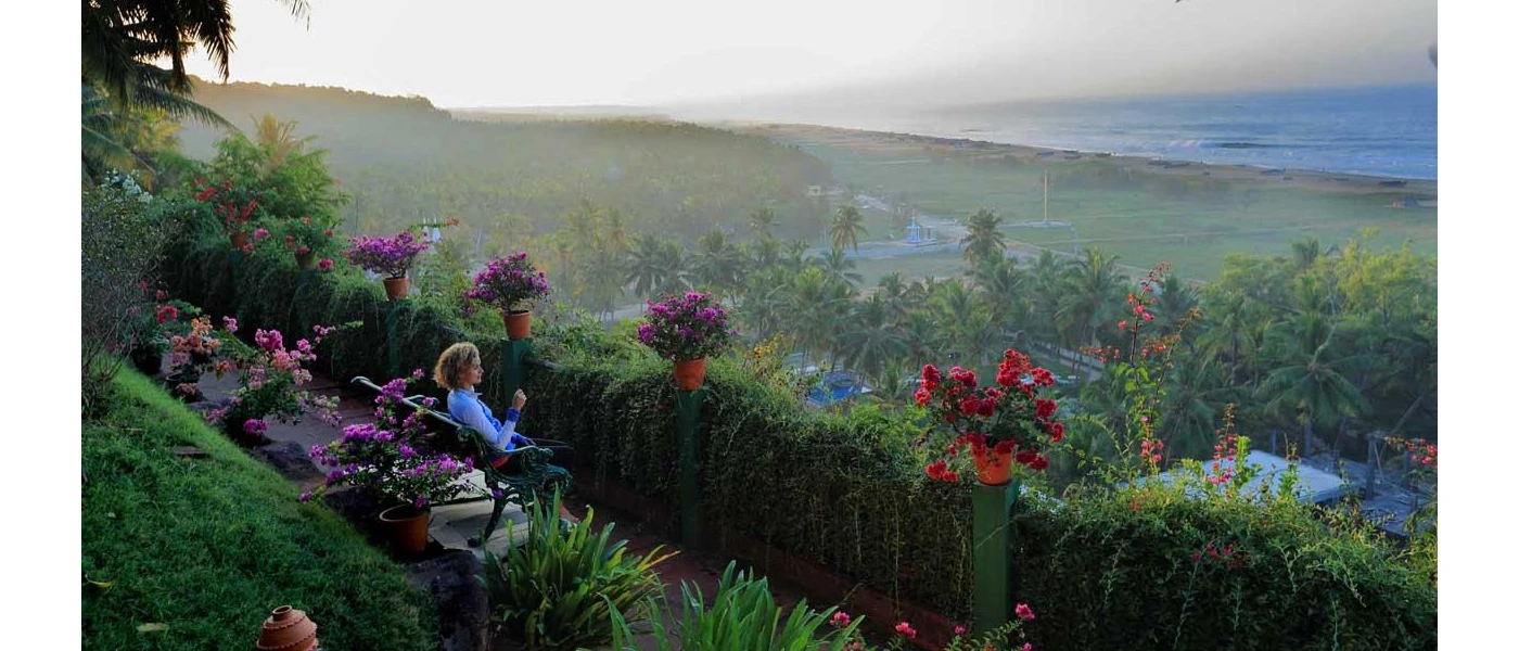 Woman sits on a bench on a terrace surrounded by tropical greenery, overlooking a misty beach and ocean