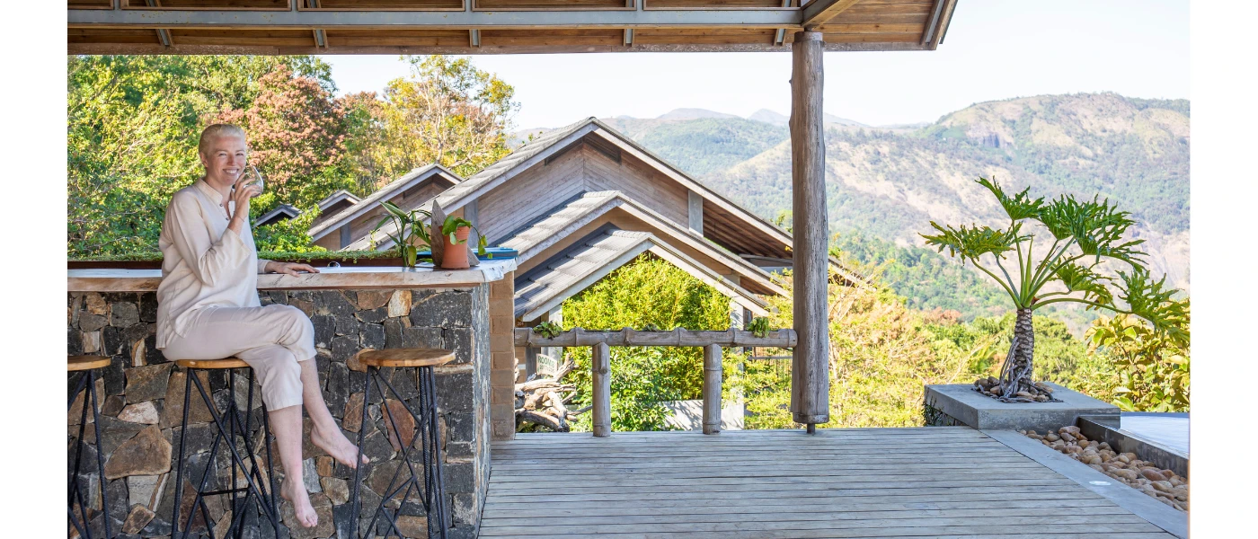 Smiling woman sips from a cup as she sits on an outdoor terrace overlooking green-cloaked mountains