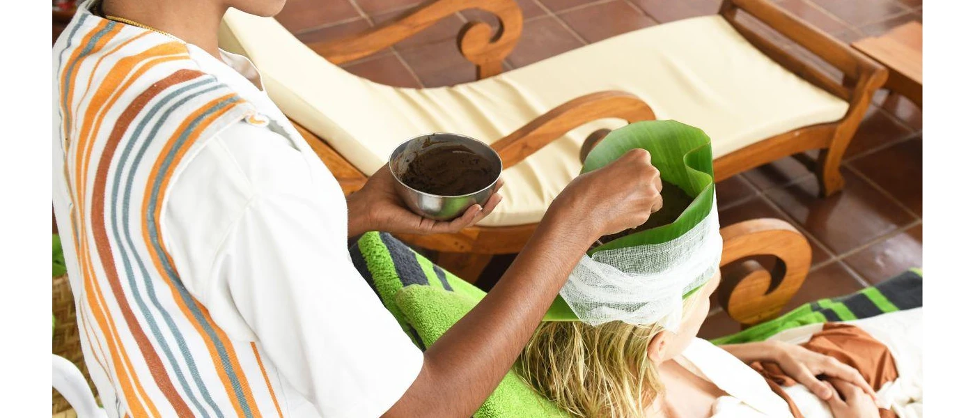 Woman sits in a head wrap consisting of large green leaves, as a therapist drips oil onto her head in a traditional Ayurveda treatment