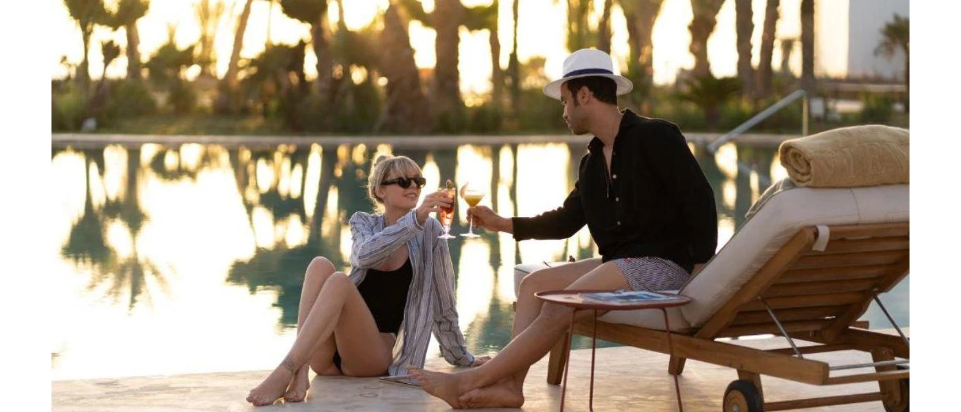 Couple toasting with a drink by the pool as the sun sets beyond the palm trees behind