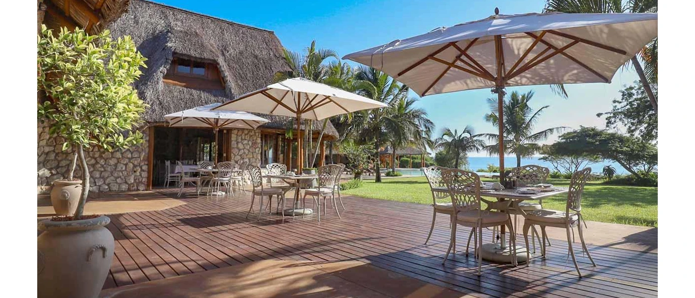 Outdoor decking with tables and chairs and large umbrellas, in front of a stone building and surrounded by trees