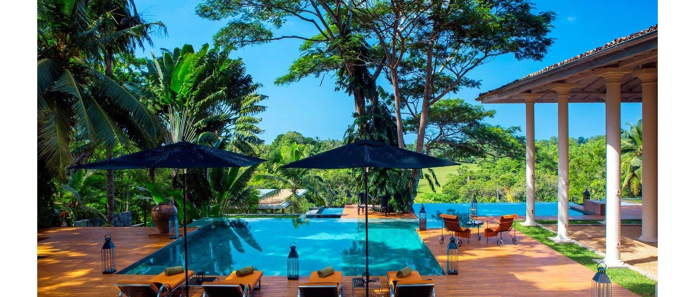 Swimming pool surrounded by black lanterns and orange cushioned loungers, next to a building with pillars and overlooking the jungle