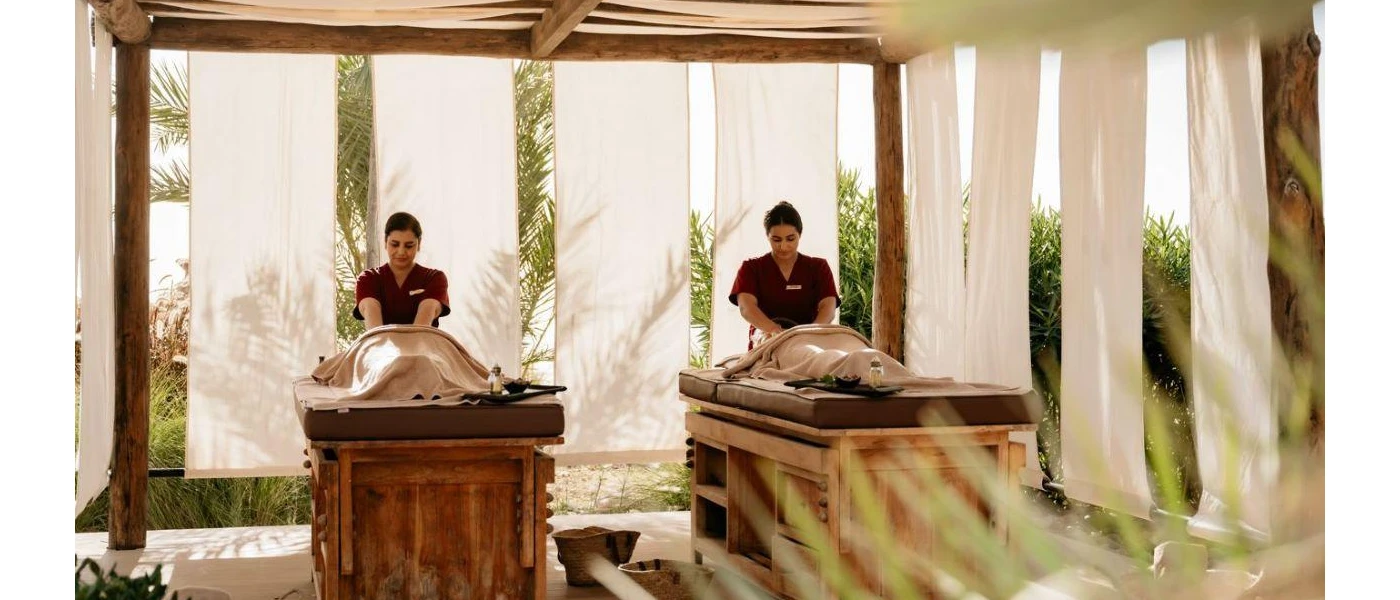Two massage therapists working on clients in an airy outdoor massage cabin, with white curtains keeping things private