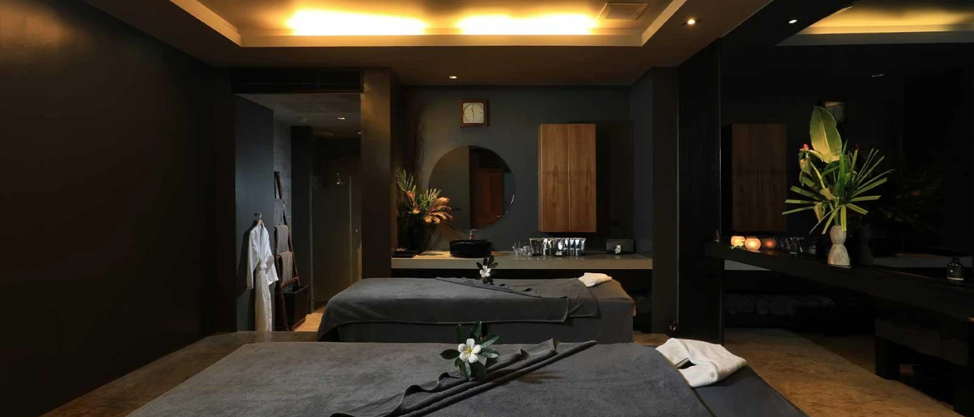 Dark-walled treatment room, with two massage beds covered in dark grey towels, hanging white robes, a circular mirror and two large vases of flowers