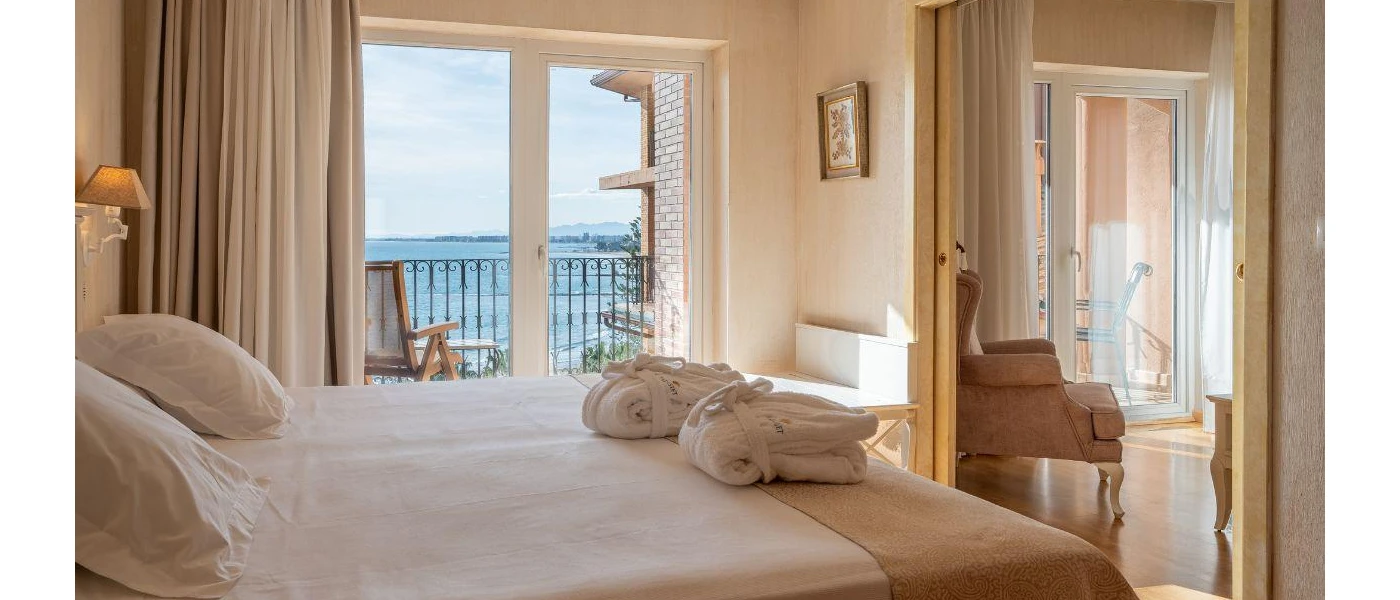 Neutral hotel bedroom with cream walls, curtains and white bedding, a wooden doorway showing an armchair, plus a balcony with ocean views