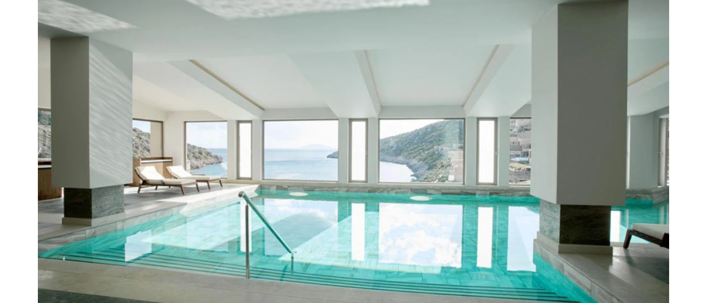 Indoor swimming pool with steps leading into it, with a terrace with two white-cushioned loungers and windows with a view of the sea and hills beyond