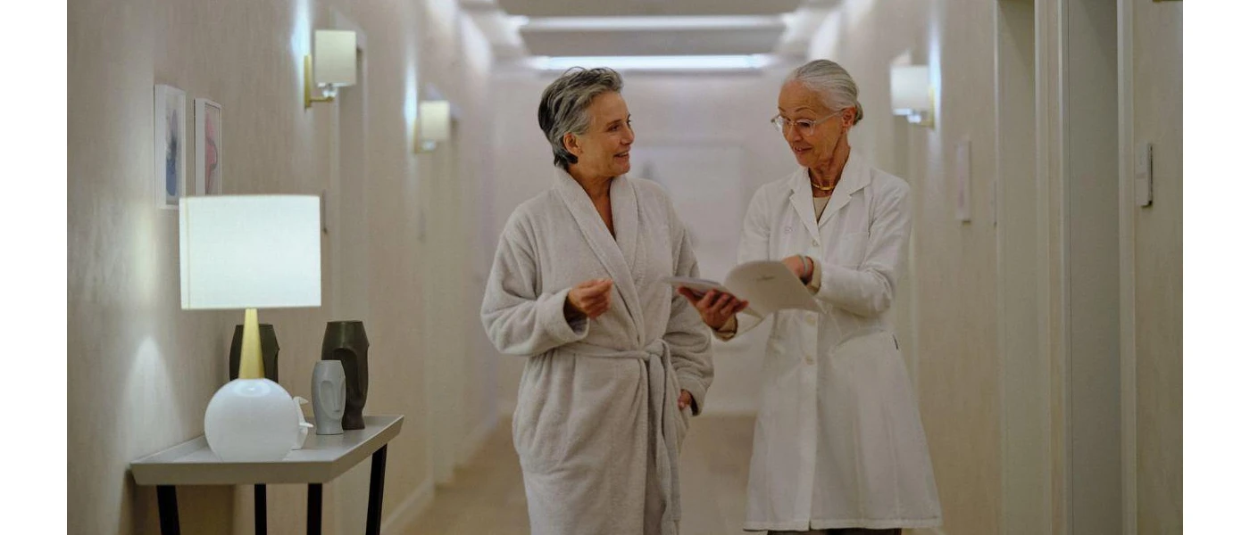 Woman in a white robe walking down a corridor with a healthcare professional in a white jacket holding a booklet open