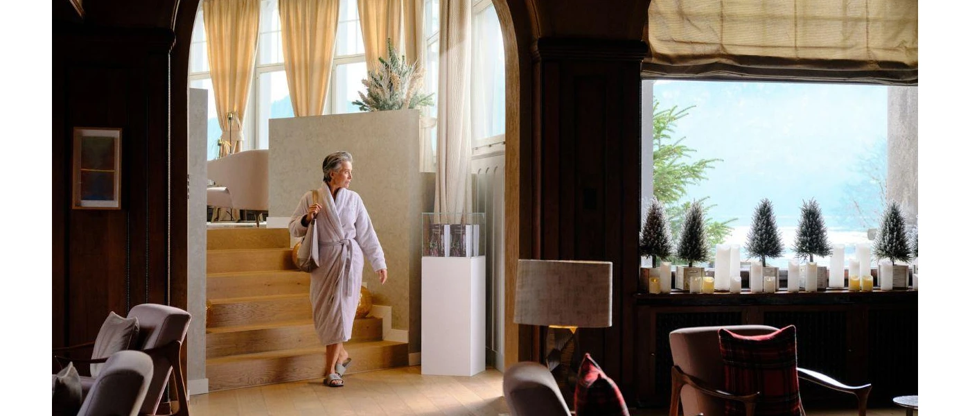 Woman in a white robe with a bag over her shoulder walking through a room with a large window and cosy seating
